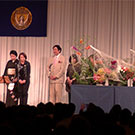 川崎景介校長に「垣間見る花」の説明を、日本語と英語でしています。約１，１００人の会員の前です。　